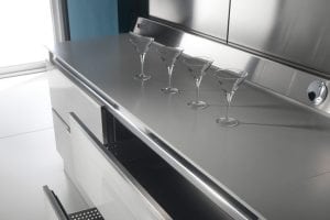 Four empty martini glasses are arranged in a diagonal line on a shiny stainless steel countertop in a modern kitchen with open drawers below.