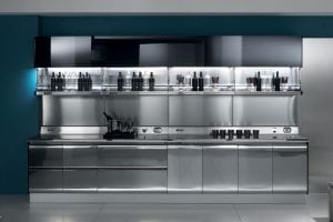 A modern kitchen with sleek, reflective stainless steel cabinets and countertops, open shelves displaying glassware and bottles, and a teal accent wall in the background. The space is brightly lit and organized.