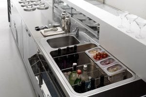 A modern kitchen counter with a double sink, ice-filled compartments holding bottles of drinks, containers of fruit, glassware, napkins, and utensils neatly arranged on a stainless steel surface.