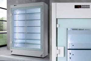 A large, glass-door commercial refrigerator with multiple empty shelves, interior LED lighting, and a digital temperature display showing 4°C. The fridge stands in a modern, minimalistic room with large windows.