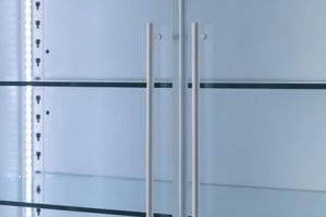 Close-up of empty glass shelves inside a modern refrigerator, with two vertical metal handles and bright interior lighting visible on the left side.