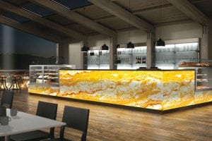 A modern café or bar with a glowing yellow and white marble-patterned counter, glass display cases with pastries, shelves with glasses and bottles, wooden floors, and large windows showing a city view at dusk.