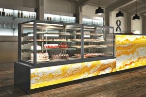 A modern bakery display case filled with assorted cakes, pastries, and desserts stands on a wooden floor. The counter has a bright yellow marble pattern. Shelves and bottles line the back wall.