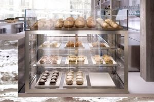 A glass display case in a bakery showcases various pastries and desserts on trays, with loaves of bread on the top shelf and a selection of cakes, eclairs, and other sweets on the lower shelves.