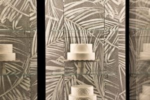 A display case with three white decorative cakes on separate glass shelves, set against a background with a large leaf pattern in shades of beige and gray.