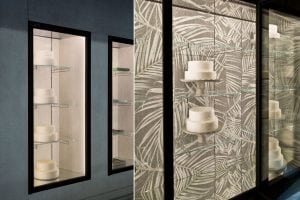 Display cases with glass shelves hold several minimalist white cakes. The case on the left has a plain background, while the right features a leafy, patterned backdrop. The cakes are unadorned and presented on stands.