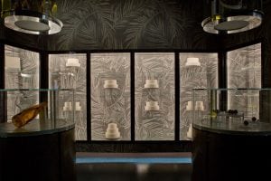 A dimly lit display room with glass cases holding objects, including a gold bottle and various small items. The back wall features lit panels with white cylindrical shapes against a patterned background.