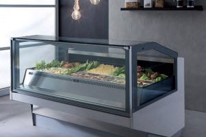 A refrigerated deli display case filled with various foods such as vegetables, salads, and meats, situated in a modern interior with hanging light bulbs and gray walls.