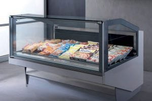 A glass display freezer filled with packaged frozen foods, including pizzas, ready meals, and desserts, is set against a gray wall in a modern retail or grocery environment.