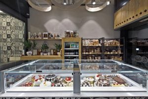 A bakery display case filled with assorted cakes and pastries. Behind the case are shelves stocked with bread, jars, and packaged goods, and the walls are decorated with patterned tiles and plants.