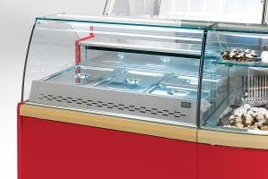 A close-up of a commercial display freezer with a glass front, featuring covered stainless steel food trays on the left and a section with assorted pastries and cookies on the right. The freezer has a red and gold exterior.