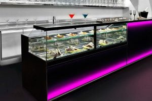 A modern glass display case with plates of assorted sandwiches and desserts is illuminated with pink LED lights. On top of the counter are three colorful cocktails and small plates, set in a stylish, contemporary bar setting.