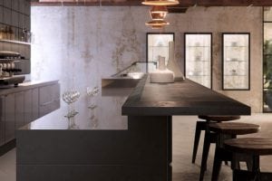 Modern kitchen with a dark countertop, three empty wine glasses, white ceramic vases, wooden stools, and glass-fronted refrigerators against a concrete wall. Copper pendant lights hang from the ceiling.