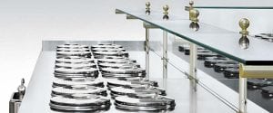 A row of shiny metal gelato containers with round lids, neatly arranged beneath a glass display shelf with brass accents in an ice cream or gelato shop.