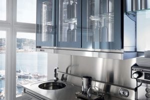 Modern kitchen with reflective stainless steel surfaces, a round sink, a coffee grinder, and glass-front cabinets containing bottles. Large windows reveal a marina with docked boats and a scenic waterfront view outside.