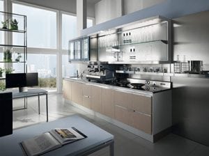 Modern kitchen with light wood cabinets, stainless steel appliances, open shelving with glassware, and large windows letting in natural light. A bookshelf and open magazine are visible in the foreground.