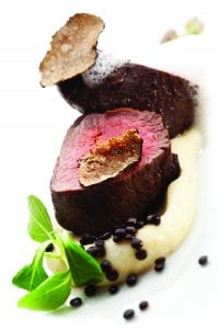 Two slices of medium-rare beef topped with truffle, served on a creamy puree, garnished with microgreens and scattered black lentils, presented on a white plate.