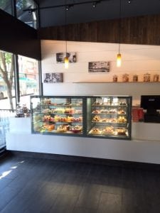A modern bakery interior with a glass display case filled with assorted pastries and desserts, white walls, hanging lights, and black-and-white photos on the wall. Large windows let in natural light from outside.