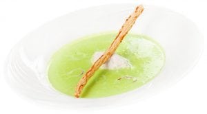 A white bowl containing creamy green soup, garnished with a dollop of white cream and topped with a long, thin, crisp bread stick.
