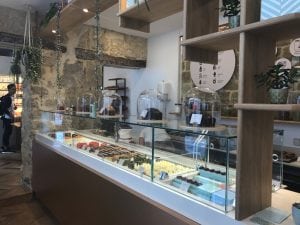 A modern chocolate shop interior with a glass display case showing various chocolates and truffles. Large glass domes cover some treats, and shelves with plants decorate the space. A person stands near the entrance in the background.