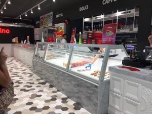 A brightly lit gelato shop with a long glass display counter filled with gelato, desserts, and pastries. Staff serve customers, and menu signs hang on the wall behind the counter. The floor has a hexagonal tile pattern.