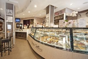 A modern pastry shop with glass display cases filled with assorted pastries and desserts. Staff work behind the counter, and there are tables and shelves with products and decorations throughout the well-lit space.