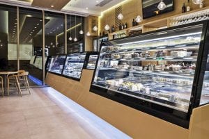 A modern bakery or café interior with glass display cases filled with assorted cakes, pastries, and desserts. The space has warm lighting, wooden accents, and a few tables and chairs for customers.