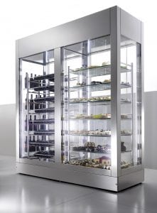 A modern, stainless steel refrigerated display case with glass doors showcases cakes, pastries, and bottles on multiple shelves in a brightly lit, minimalist setting.