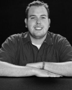 A young man with short hair and a beard smiles at the camera, sitting at a table with his arms crossed. He is wearing a dark button-up shirt and a wristband, with a plain black background behind him.
