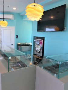 A modern self-serve frozen yogurt shop interior with empty topping containers, a soft-serve machine, a wall-mounted TV, a metal sink, bright turquoise walls, and yellow pendant lights.