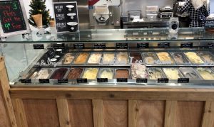 A display case with various flavors of ice cream in tubs, labeled with small black signs. Behind the counter are ice cream machines, utensils, and a small menu board. A small Christmas tree decorates the left side.