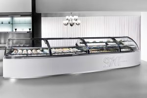 A modern, curved gelato display case filled with various flavors of gelato sits in a sleek, minimalist shop with white walls, a chandelier, and glass shelves in the background.