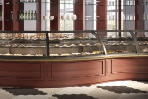 A sleek bakery display case showcases assorted pastries and desserts on glass shelves, set against wooden cabinetry and wall shelving with bottles, cups, and plates. Hexagonal tile flooring is visible in the foreground.