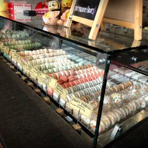 A glass display case filled with colorful macarons in various flavors, each labeled with signs. Plush toys and a chalkboard menu are visible on the counter above the case.