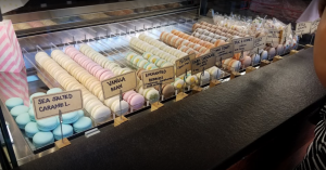 A display case of colorful macarons in various flavors such as sea salted caramel, vanilla bean, enchanted berries, lemon, dark chocolate, cinnamon toast, and milk & honey, each labeled with a sign.