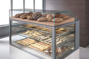 A glass bakery display case with assorted loaves of bread on top and shelves filled with pastries, cookies, and desserts inside, all neatly arranged in a modern, well-lit setting.