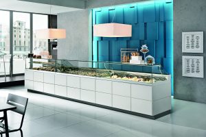 A modern gelato shop interior with a long glass display case filled with colorful gelato, a blue accent wall, two pendant lights, and tables and chairs in the foreground.