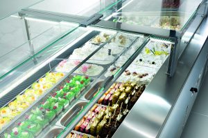 A display case filled with various colorful gelato and ice cream flavors, topped with fruits and decorations, in a well-lit, modern shop setting.