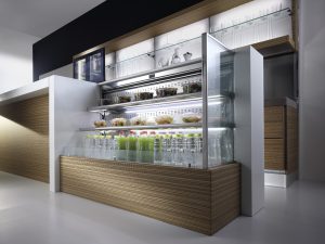 A modern refrigerated display case holds bottled juices, water, salads, and fresh produce. The unit has wood paneling, glass shelves, and is part of a sleek, contemporary kitchen setup.