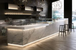 A modern café with a marble counter, glass display case, bar stools, and geometric pendant lights. Shelves with glasses and a view of a river through large windows are visible in the background.