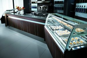 A modern bakery or café interior with glass display cases filled with pastries, a countertop with fruit arrangements, and shelves lined with bottles in the background. The space is stylish and clean with dark wood accents.