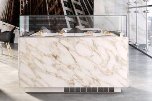 A modern marble-fronted dessert display case with pastries inside, set in a sleek, glass-walled room with city buildings visible through the windows.