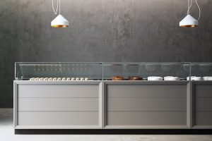 A modern bakery display counter with pastries and cakes behind glass, set against a gray concrete wall. Two white pendant lights hang from the ceiling above the counter.