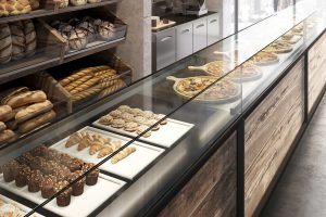 A bakery display case with trays of muffins, croissants, and pastries, alongside several pizzas on wooden boards. Shelves behind the case hold loaves and baguettes. The interior features wood and glass elements.