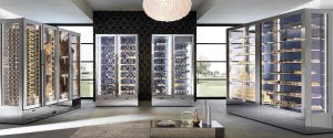 A modern, elegant room with glass display refrigerators showcasing wine, desserts, cakes, and cured meats. Large windows and a central chandelier add brightness to the contemporary space.