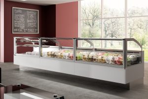 A modern ice cream parlor with a long glass display case filled with colorful gelato tubs. The room has large windows, red walls, and a chalkboard menu on the wall. Natural light brightens the clean interior.