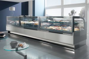 A modern bakery display case filled with assorted pastries and cakes stands in a bright, stylish shop with large windows; a glass table with a coffee cup is in the foreground.