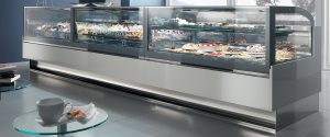 A modern glass display case in a bakery filled with various cakes, pastries, and desserts. In the foreground, a round glass table holds a cup of coffee and a plate with a pastry.