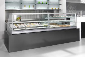 A modern display case filled with various pastries and desserts sits on a sleek black counter in a minimalist, brightly lit shop with decorative glassware on shelves in the background.