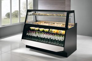 A refrigerated display case filled with assorted pastries on the top shelf, salads on the middle shelf, and bottles of water on the bottom shelf, set in a modern, bright room with large windows.